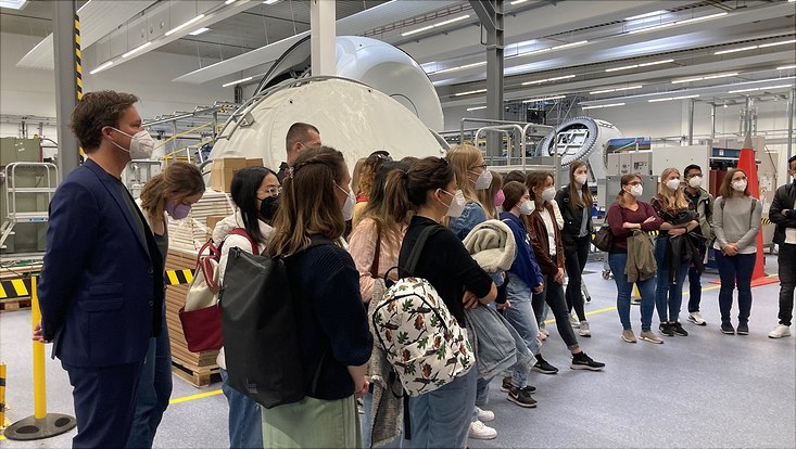 Prof. Busch und Studierende hören einem Vortrag in einer Industriehalle zu